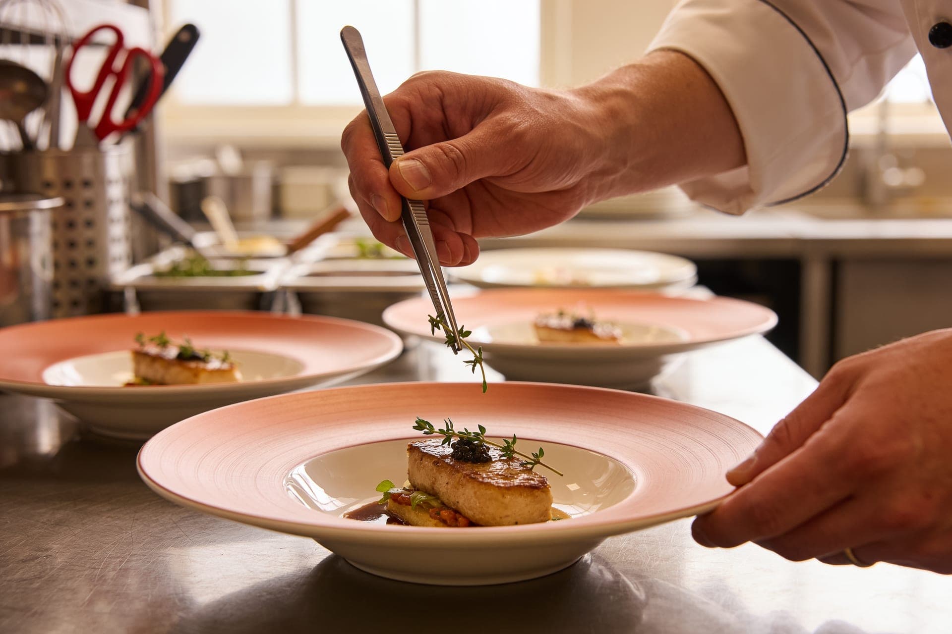 A chef plating a course with tweezers at the pass, garnishing with herbs